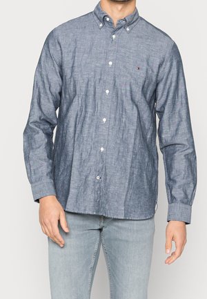 Muž nosící modrou chambray košili s dlouhým rukávem, zapnutou na knoflíky, s malým logem na hrudi, v kombinaci se světle modrými džínami.