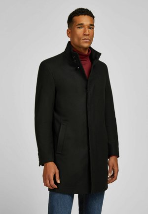 Schwarzer Wollmantel mit hohem Kragen, Knopfdetails, vorderen Taschen und geradem Schnitt. Trägt man über einem bordeauxfarbenen Rollkragenpullover und blauen Jeans.