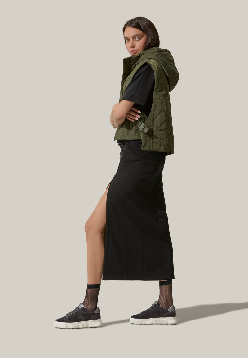 Gilet imbottito verde oliva con cappuccio, t-shirt nera, gonna nera con spacco laterale e sneakers grigie con suole bianche. Calze nere testurizzate visibili.