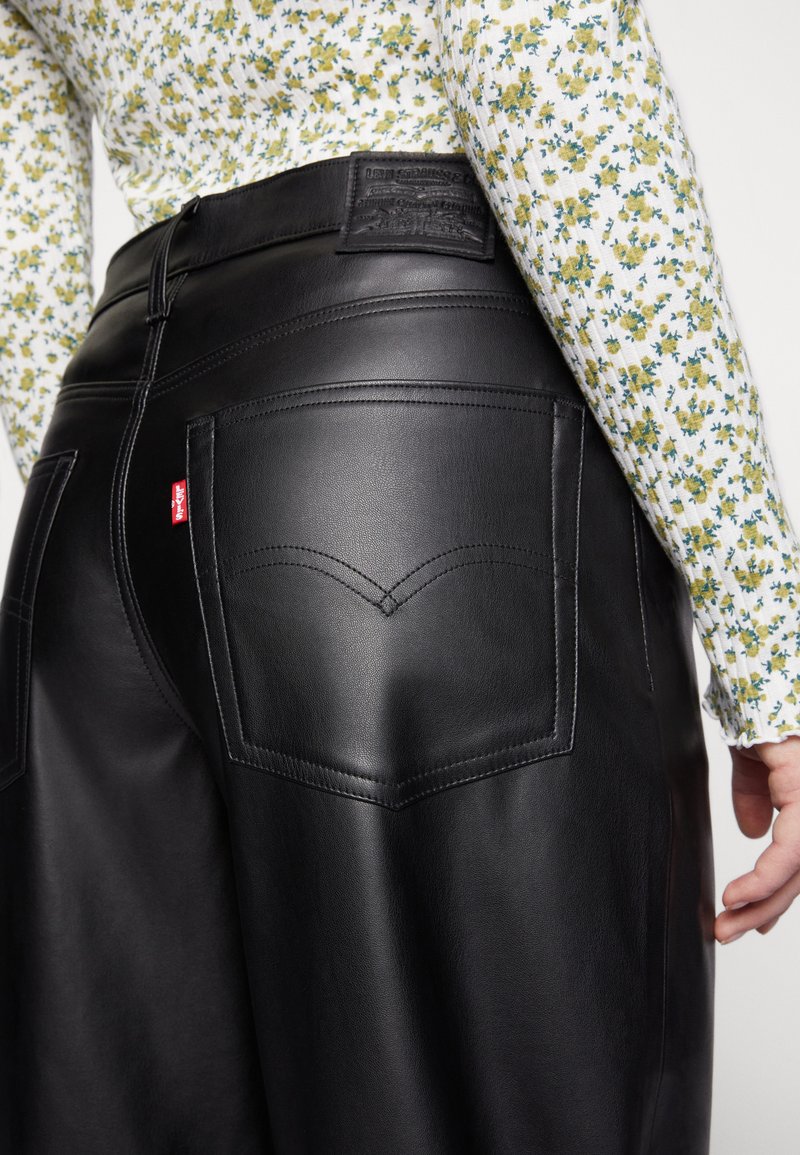 Close-up van een persoon die zwarte leren Levi's broeken met een hoge taille draagt, gecombineerd met een bloemen top met lange mouwen met gele en groene bloemen.