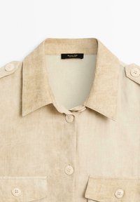 Beige Leinenhemd mit Kragen, ausgestattet mit zwei vorderen Taschen und Knopfakzenten. Weiche Textur mit einem subtilen Glanz und detaillierten Nähten.