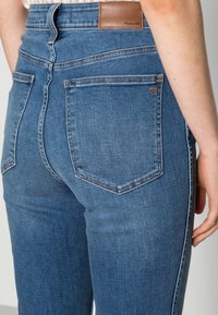 Högmidjade blå denimjeans med en brun läderlapp, subtila sömnadsdetaljer och klassiska bakfickor.