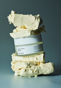 Hvit rund krukke med ansiktskrem, omgitt av en kremet, beige substans med tekstur som likner sheasmør. Etiketten viser produktnavn og prosentandel naturlige ingredienser.