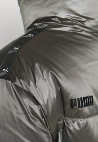 Mörkgrå pufferjacka med glansig yta, med en svart logotyp och mönstrad rem längs axeln. Slät textur, vadderad design.