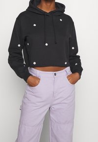 Schwarzer, kurz geschnittener Hoodie aus Baumwolle mit weißer Gänseblümchen-Stickerei, die über das gesamte Kleidungsstück verteilt ist. Kombiniert mit hellvioletten Cargo-Hosen.