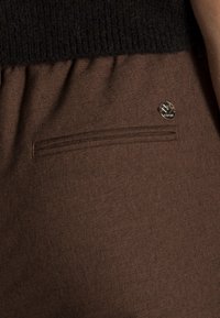 Brunt tyg med en texturerad yta, med ett svart stickat midjeband och en liten dekorativ metallknapp på bakfickan.