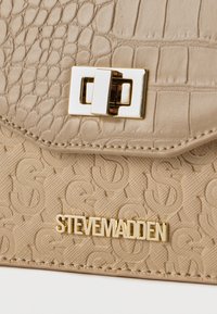 Μπεζ υφασμάτινη τσάντα με ανάγλυφα μοτίβα S, χρυσό ορθογώνιο κλείστρο με περιστροφικό κούμπωμα και χρυσή πλακέτα με το λογότυπο STEVEMADDEN.