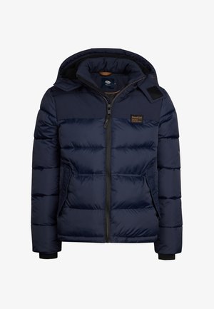 Chaqueta acolchada azul marino con cremallera frontal, capucha ajustable y bolsillos laterales, combinada con jeans azules y zapatillas deportivas negro/blanco.