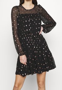 Vestido negro con mangas largas y transparentes, adornado con lunares en oro rosa, una cintura fruncida y una falda fluida. La tela texturizada añade dimensión.