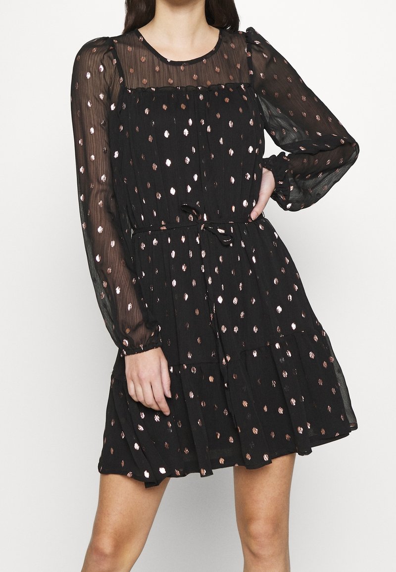 Vestido negro con mangas largas y transparentes, adornado con lunares en oro rosa, una cintura fruncida y una falda fluida. La tela texturizada añade dimensión.