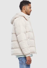 Pufferjakke i lys beige med lynlås foran, hætte og ribbede manchetter. Lavet af isolerende materiale med quiltet design og sidelommer.