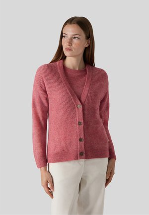 Kvinde iført en struktureret pink knappet cardigan over en matchende pink top og hvide bukser, kigger let mod venstre mod en grå baggrund.