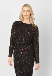 Zwarte lange mouw jurk bedekt met rode en zilveren pailletten, strakke silhouet, ronde hals, met transparant materiaal bij de mouwen en een glanzende textuur.