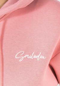 Sudadera rosa con una textura suave, que presenta un logotipo en cursiva blanco de "Smilodon" en el pecho y una capucha espaciosa.