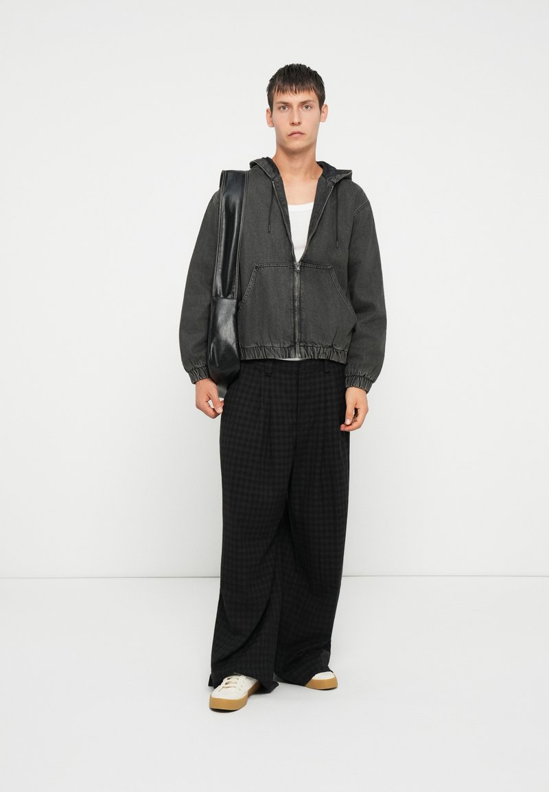 Chaqueta de mezclilla negra con capucha, dobladillo elástico y bolsillos frontales. Combinada con pantalones sueltos de cuadros negros y zapatillas beige.