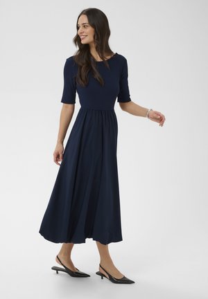 Femme portant une robe midi bleu marine à manches mi-longues et des escarpins slingback noirs à petit talon, se retournant légèrement et souriant sur un fond uni.