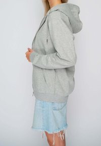 Šedá mikina s zipem, měkkou texturou, nastavitelnou kapucí a předními kapsami, kombinována s roztrhanou džínovou sukní.