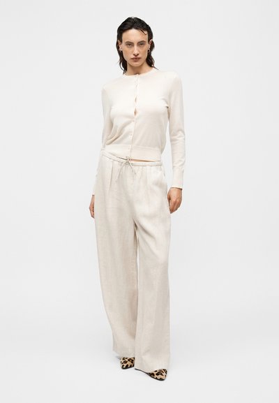 Ανοιχτό μπεζ ζακέτα με στρογγυλή λαιμόκοψη και κούμπωμα κουμπιών, συνδυασμένη με φαρδιά μπεζ λινά παντελόνια και τακούνια με λεοπάρ σχέδιο. Ομαλή υφή συνολικά.