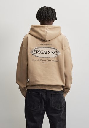 Person med kort snoet hår iført en beige hættetrøje med teksten "PEGADOR Time to Pursue Your Dreams" på ryggen og sorte jeans.