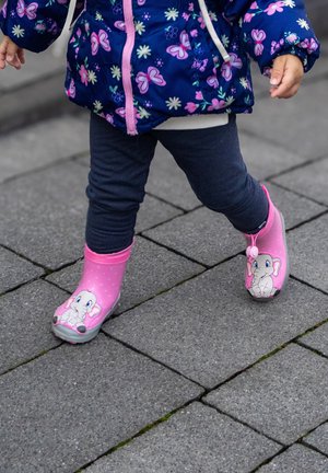 Kind in marineblauen Leggings und einer Blumenjacke, das auf dem Bürgersteig läuft und rosa Stiefel mit cartoonartigen Elefantengesichtern trägt.