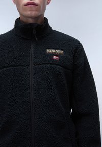 Black fleece jacket with a textured surface, high collar, and front zipper. Features a logo patch with text and a small flag detail.