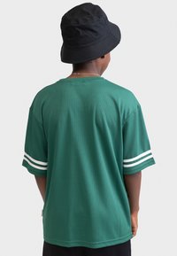 Grünes Mesh-T-Shirt mit weißen Streifen an den Ärmeln, Rundhalsausschnitt, lockere Passform und glatte Textur, kombiniert mit einem schwarzen Bucket Hat.