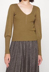 Olivengrünes, geripptes Strickoberteil mit tiefem V-Ausschnitt, vorderem Knopfdetail und Ballonärmeln, kombiniert mit einem silbernen, glitzernden Rock.