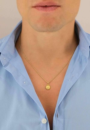 Collier en or avec un pendentif rond, présentant un design complexe, porté sur une chemise col bleu clair, montrant les détails et la texture du col.