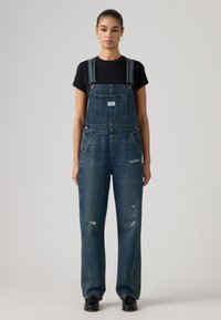 Νεαρή γυναίκα όρθια, φορώντας σκούρο τζιν σαλοπέτα με φθαρμένες λεπτομέρειες, μαύρο μπλουζάκι, μαύρα παπούτσια και κρίκους στα αυτιά, σε γκρι φόντο.