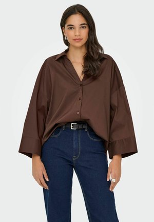 Femme aux longs cheveux foncés portant une chemise ample marron à boutons, un jean bleu foncé, une ceinture noire, des boucles d'oreilles argentées et une bague en argent, se tenant devant un fond blanc.