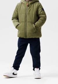 Grøn polstret jakke med hætte og lynlås, kombineret med mørkeblå teksturerede bukser og hvide sneakers med sorte detaljer.