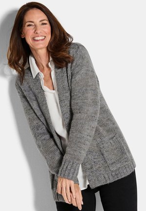 Lächelnde Frau mit braunen Haaren, die einen grauen Strickcardigan über einem weißen Shirt und einer schwarzen Hose trägt, vor einem schlichten Hintergrund stehend.