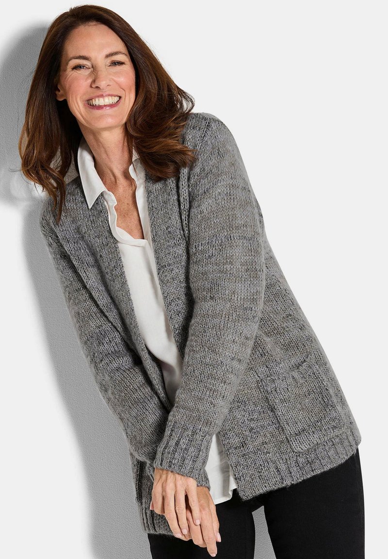 Lächelnde Frau mit braunen Haaren, die einen grauen Strickcardigan über einem weißen Shirt und einer schwarzen Hose trägt, vor einem schlichten Hintergrund stehend.