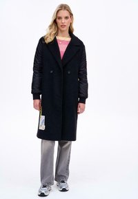 Mladá žena ve dlouhém černém kabátě s prošívanými rukávy, růžovém svetru, šedých džínách a stříbrných teniskách, stojící před jednobarevným bílým pozadím.