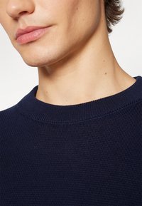 Maglione lavorato a maglia blu navy con collo rotondo, caratterizzato da un tessuto testurizzato e un colletto a coste, con attenzione alla zona superiore del torace e del collo.