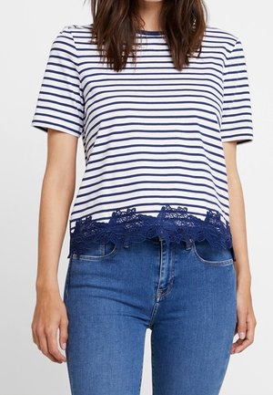 Vrouw draagt een wit marine gestreept shirt met korte mouwen en een marineblauw kanten randje aan de zoom, gecombineerd met blauwe spijkerbroek.