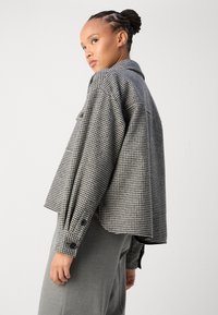 Black and white houndstooth pattern shirt jacket with a loose fit, pointed collar, and buttoned cuffs, paired with grey trousers.