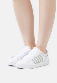 Sneakers bianchi con accenti grigi, caratterizzati da un materiale morbido e un design a punta tonda. Chiusura con lacci e tre strisce sul lato.