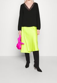 Femme portant un chemisier noir orné de dentelle, une jupe en satin jaune néon, des bottes à talons hautes noires, tenant un petit sac à main rose vif noué.