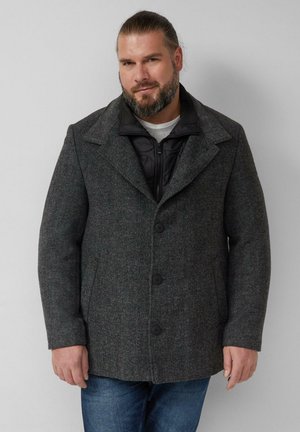 Man met baard die een donkergrijze wollen jas met knopen draagt over een zwart doorgestikt jack en een blauwe spijkerbroek, staand tegen een effen lichte muur.