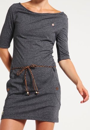 Kobieta ubrana w ciemnoszarą sukienkę do kolan z rękawami do łokci i zaplecionym brązowym paskiem przewiązanym w talii.
