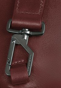 Detalle de bolso de cuero burdeos con un clip metálico de acabado brillante y nombre de la marca en relieve, mostrando costuras y matices de textura.