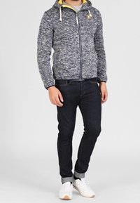 Marinblå stickad zip-jacka med gula detaljer och dragsnören, tillsammans med mörka jeans och vita sneakers.