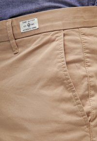 Pantaloni chino beige con una texture liscia, dotati di tasca standard, passanti per la cintura e un'etichetta che mostra il nome del marchio e l'anno di fondazione.