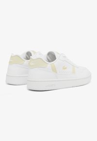 Zapatillas de cuero blancas con una textura suave, que presentan detalles en amarillo claro en el talón y el costado, y una marca en relieve en la suela de goma.