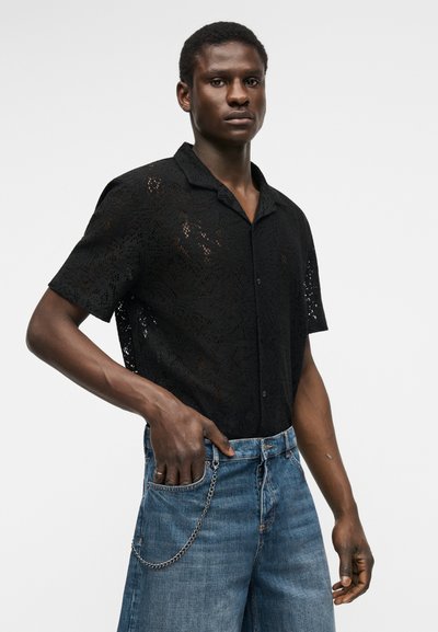 Άνδρας με μαύρο πουκάμισο από δαντέλα με κοντά μανίκια και μπλε τζιν με αλυσίδα, στέκεται μπροστά σε απλό ανοιχτό φόντο.