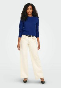 Sinine kudumiskampsun pikade varrukatega, koos kreemjast laiade pükste ja mustade teravate kingadega. Omab vööd ja lokkis juukseid.