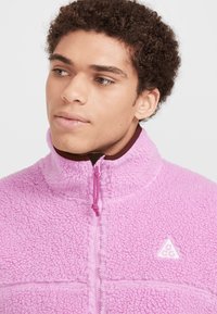 Rosa fleecejacka med dragkedja i kragen, texturerad tyg, liten logotyp på vänster bröst och kontrasterande färg på insidan. Anmärkningsvärt dragkedjedetalj på kragen.