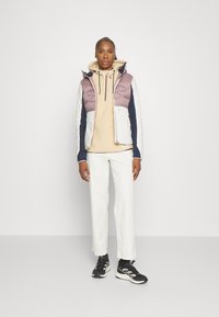 Polstret vest i lys lilla, hvit og marineblå; kombinert med en beige hettegenser, hvite bukser og svarte joggesko; har glidelåslukking og lommer.