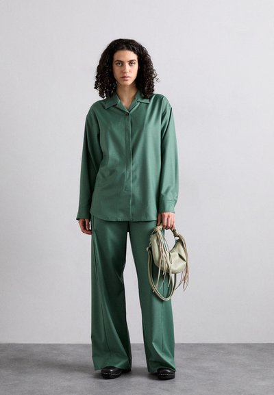 Una camicia verde a maniche lunghe con colletto, abbinata a pantaloni a gamba larga. Il modello tiene una piccola borsa a tracolla verde chiaro con dettagli testurizzati.